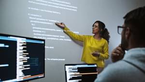 Weibliche Mentorin hält einen Vortrag und steht vor einer Wand auf dem ein Beamer bläuliche Codes projiziert. 